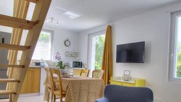A cozy room with a wooden staircase, a dining area with a table and chairs, a TV mounted on the wall, and a sofa with a small table.