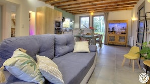 A modern living room with a grey sofa, wooden ceiling beams, a dining area, and a kitchen with wooden cabinets. - la Ferme aux Colombages (MARBOZ)