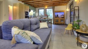A modern living room with a grey sofa, wooden ceiling beams, a kitchen in the background, and a dining area with a yellow chair. - la Ferme aux Colombages (MARBOZ)