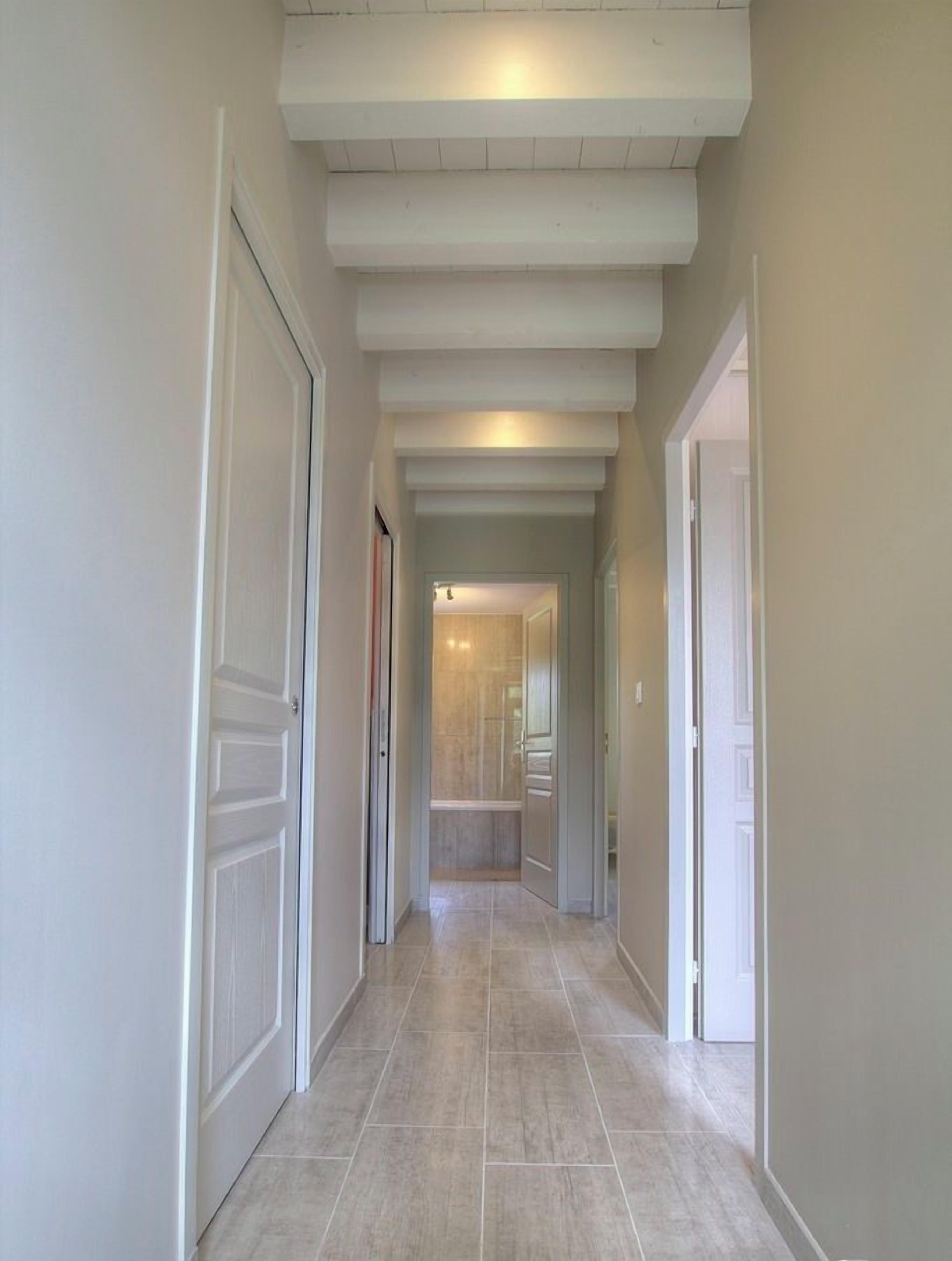 Un pasillo con puertas blancas, piso de baldosas y techo con vigas a la vista.