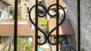 Ein Balkon mit herzförmiger Metallreling, der auf ein Wohngebiet mit Steinhäusern und einen grünen Hügel blickt.