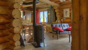 A log cabin interior with a central wood stove, wooden walls, a dining area, and a leather sofa. - Chalet La Fuste Monts Jura (LELEX)