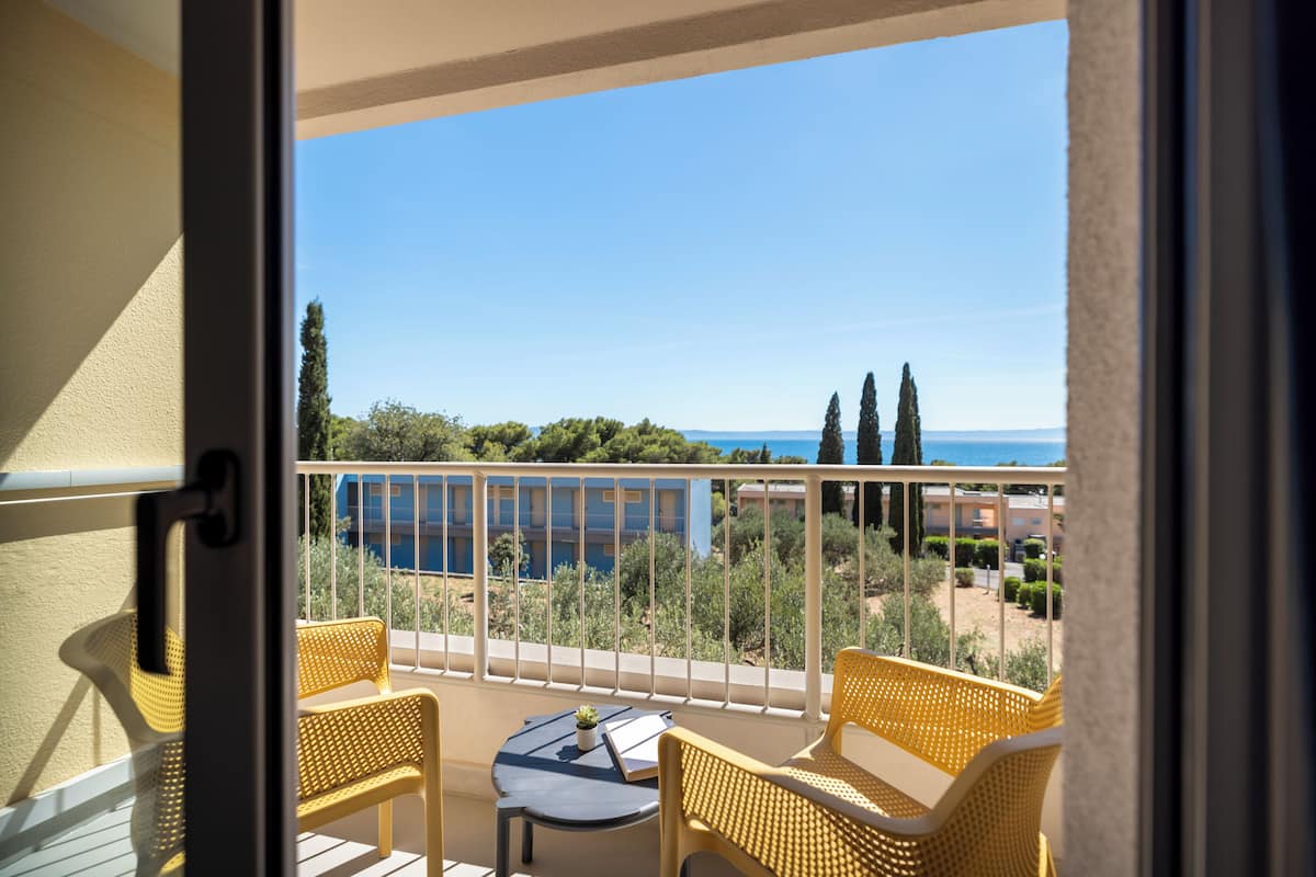 Un balcon avec deux fauteuils en osier et une petite table, donnant sur une zone côtière avec des bâtiments et des arbres.