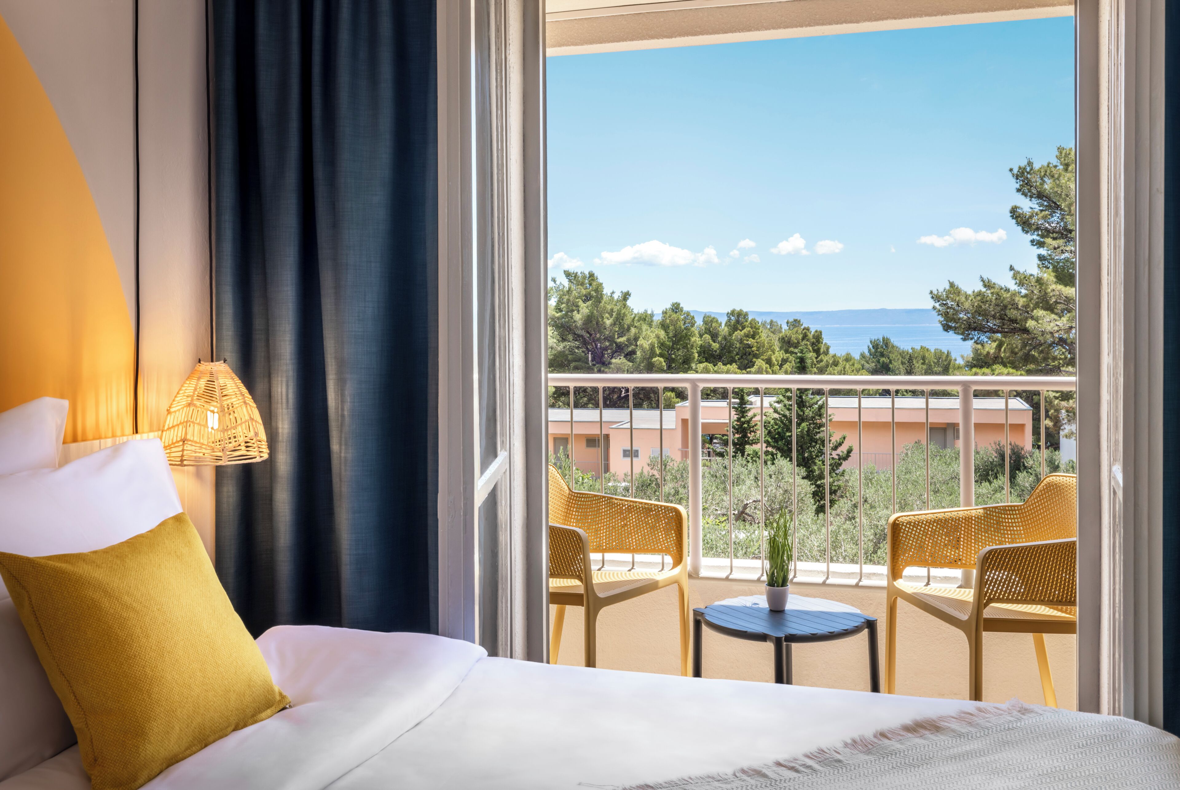 A hotel room with a bed, a lamp, a view of the sea, and two wicker chairs on a balcony.