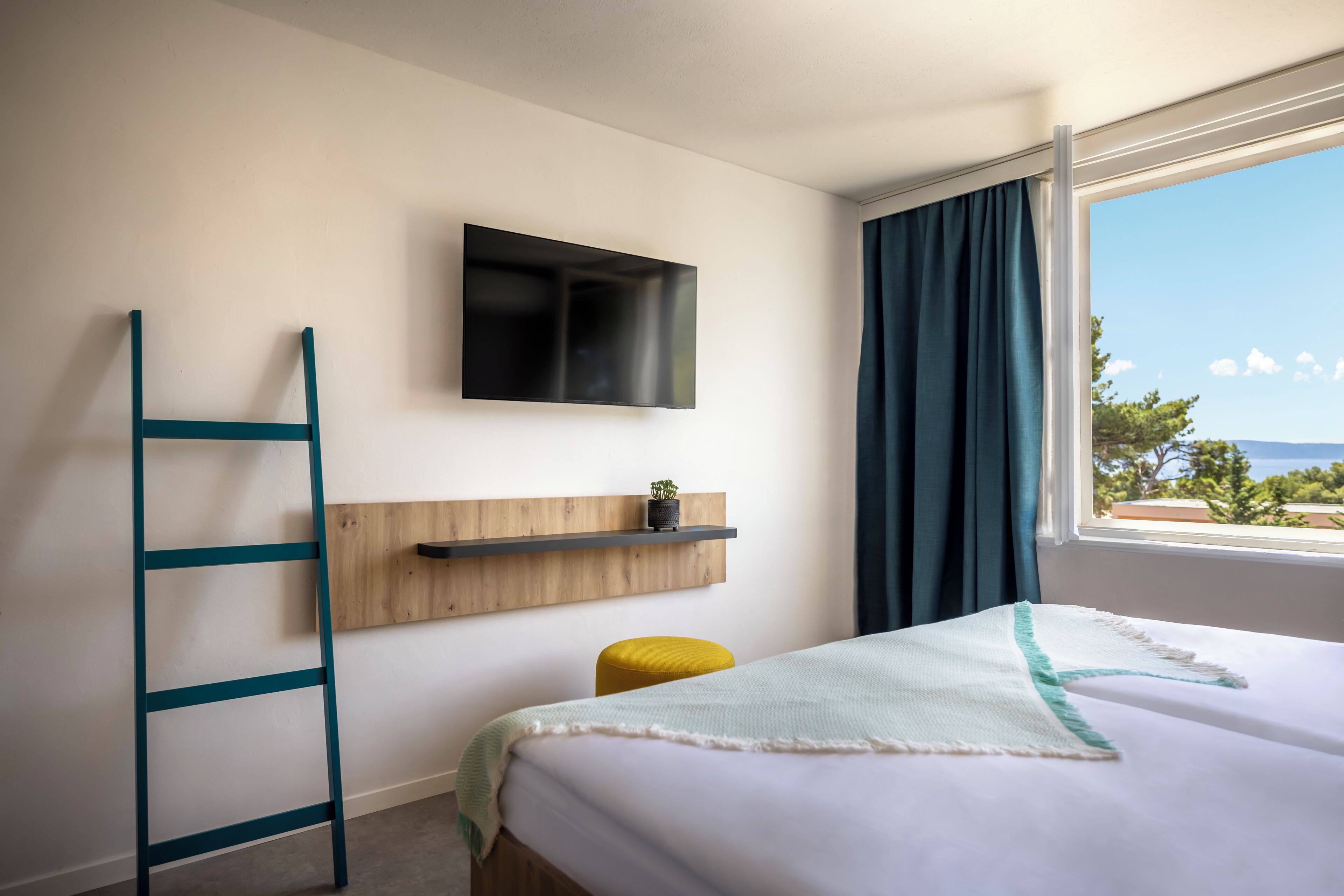 A hotel room with a bed, a TV mounted on the wall, a wooden shelf with a plant, a yellow stool, and a window with curtains.