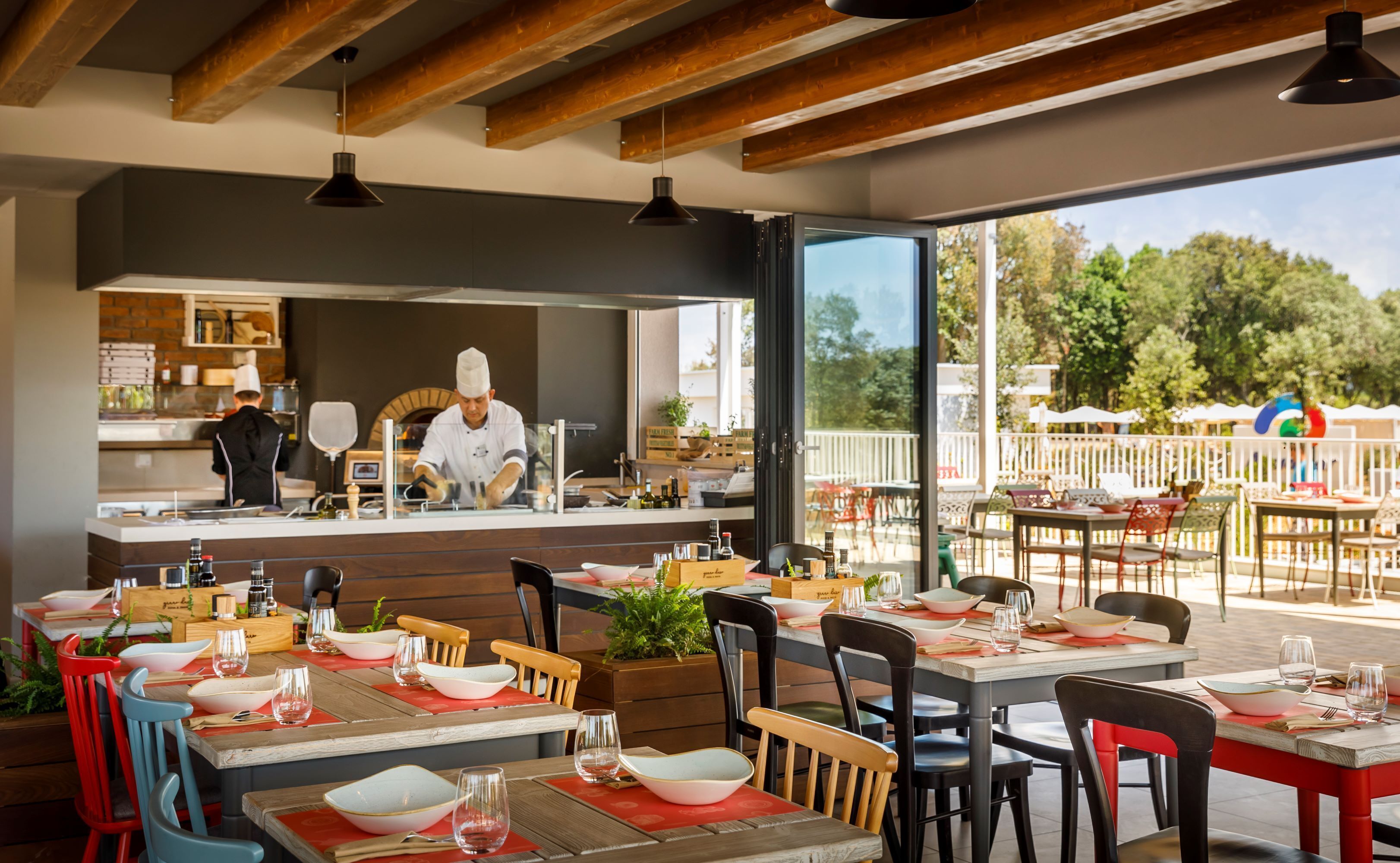 Un espacio exterior para comer con mesas y sillas de madera, un chef preparando comida y vistas a árboles y una cerca blanca.