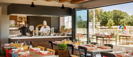 Un espacio exterior para comer con mesas y sillas de madera, un chef preparando comida y vistas a árboles y una cerca blanca.