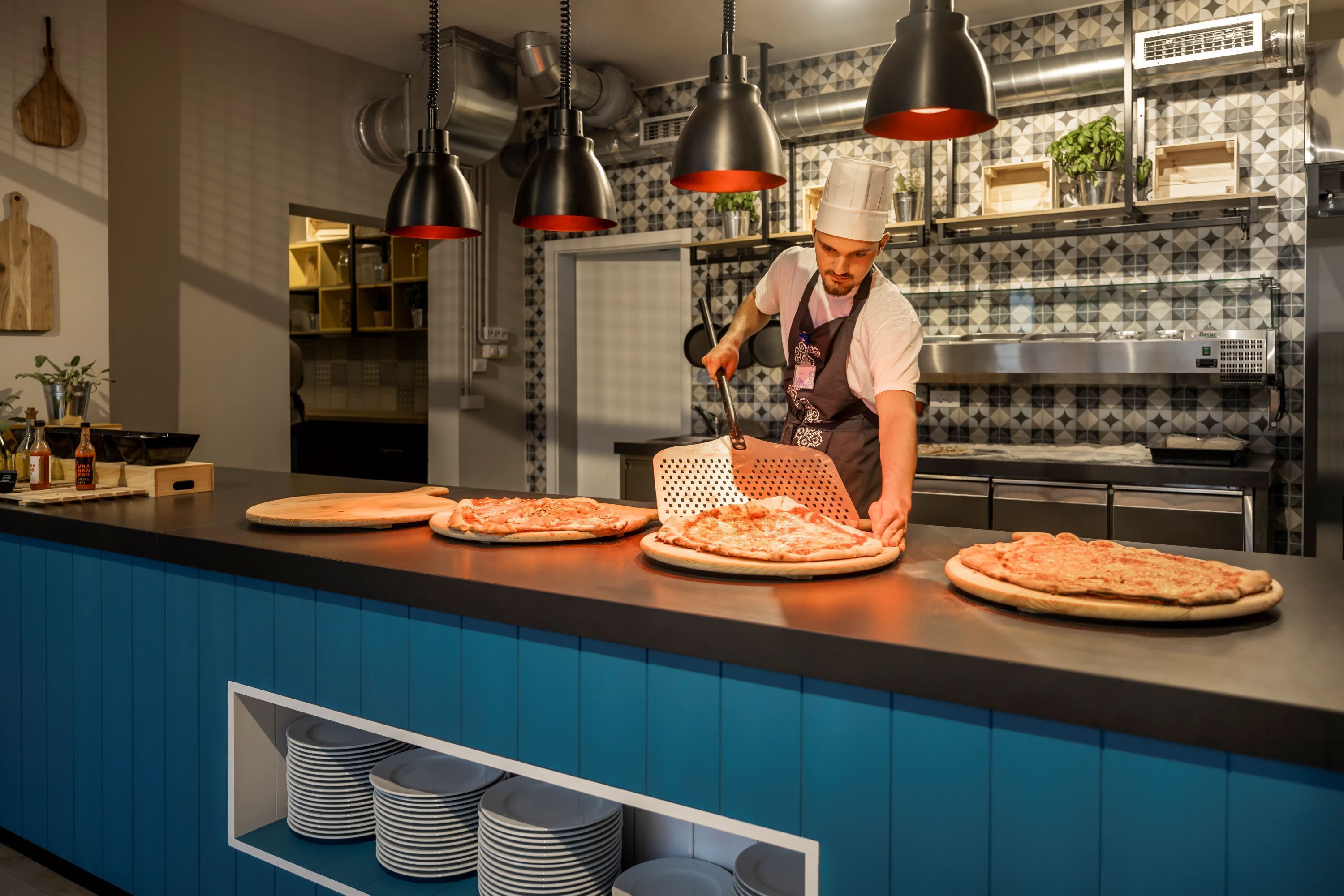 A chef preparing pizzas in a modern kitchen with blue cabinetry and black pendant lights.