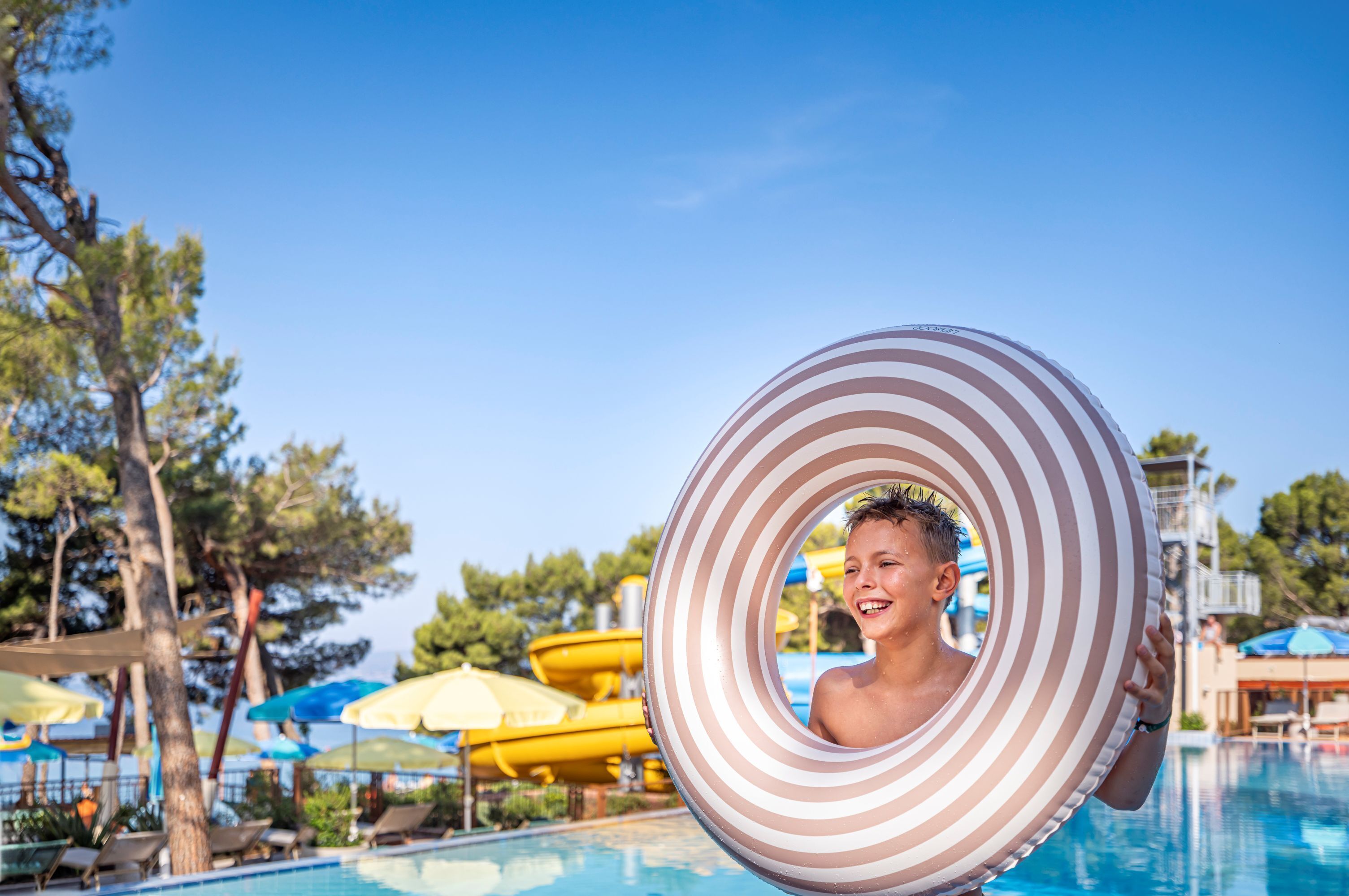 A swimming pool with lounge chairs and umbrellas, a water slide, and trees in the background.