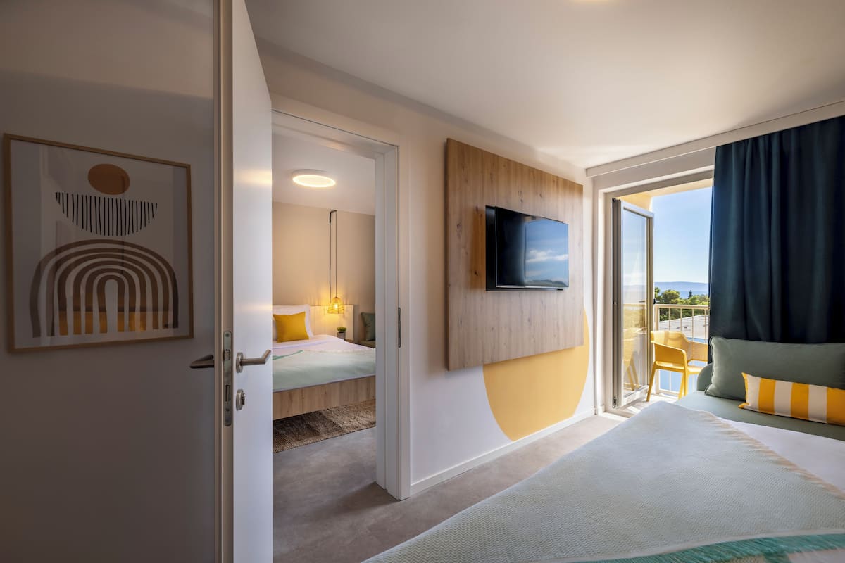 Une chambre d’hôtel moderne équipée d’un lit, d’un téléviseur fixé au mur, d’un balcon avec vue et d’une porte donnant sur une autre chambre.