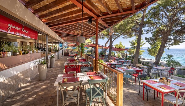 Un restaurante al aire libre con mesas y sillas, vista al mar y un edificio con un letrero rojo.