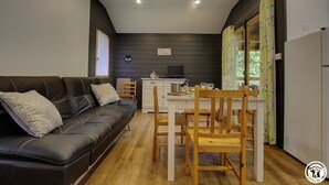 Un espacio habitable compacto con un sofá de cuero negro, una mesa de comedor de madera con sillas, una pequeña cocina y un ventanal con cortinas florales.