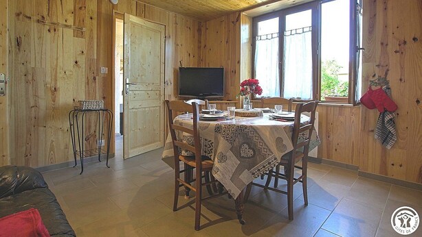 Um quarto com painéis de madeira, uma mesa de jantar posta para quatro pessoas, uma televisão e uma janela com cortinas.
