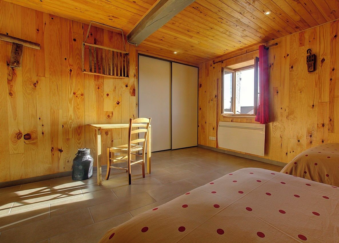 A wooden room with a bed, a table, and a chair. There is a window with curtains, and a radiator on the wall.
