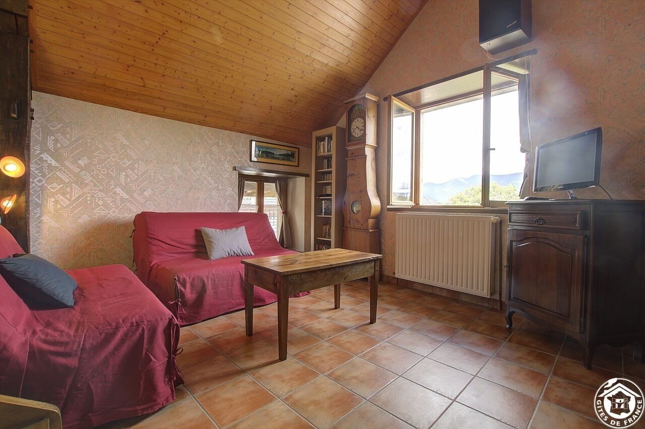 Ein gemütliches Zimmer mit Holzbalkendecke, einem roten Sofa, einem Holztisch, einem Fernseher und einem Fenster mit Blick auf die Berge.