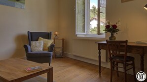 A cozy room with a wooden floor, a blue armchair, a small table with a book, a wooden desk with a chair, a window with a view of houses, and a vase with flowers on the desk. - la Grande Fontaine - Ceyzeriat (CEYZERIAT)