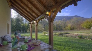 A wooden pergola with a table set for a meal, overlooking a grassy area with trees and mountains in the background. - Les Epées - Conzieu - Gîte (CONZIEU)