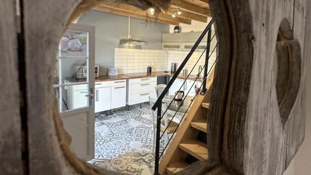 A wooden structure with a circular window revealing a kitchen with white cabinets, a patterned rug, and a staircase.