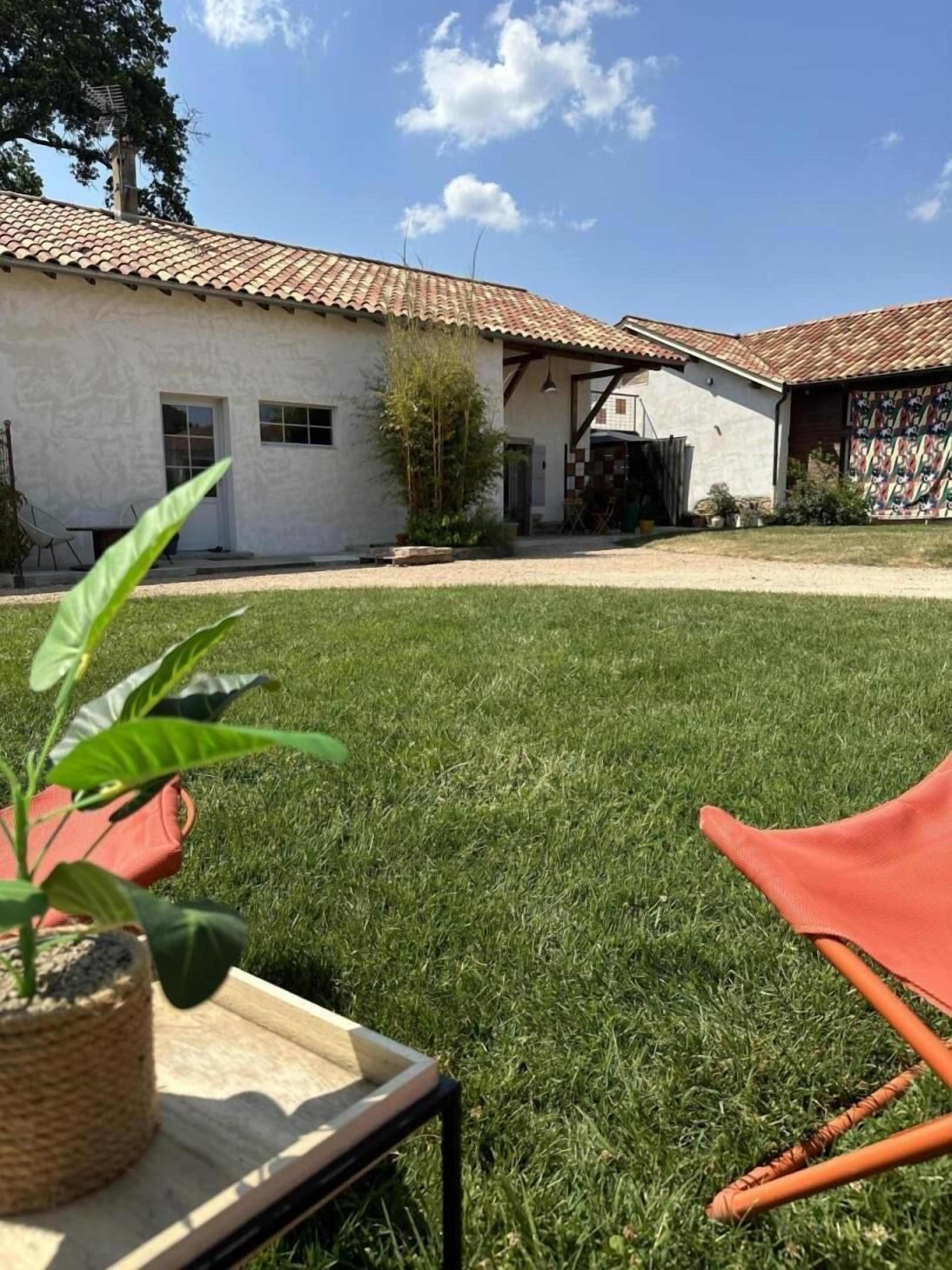 A grassy yard with a building that has a tiled roof and a door. There is a chair and a plant in the foreground.