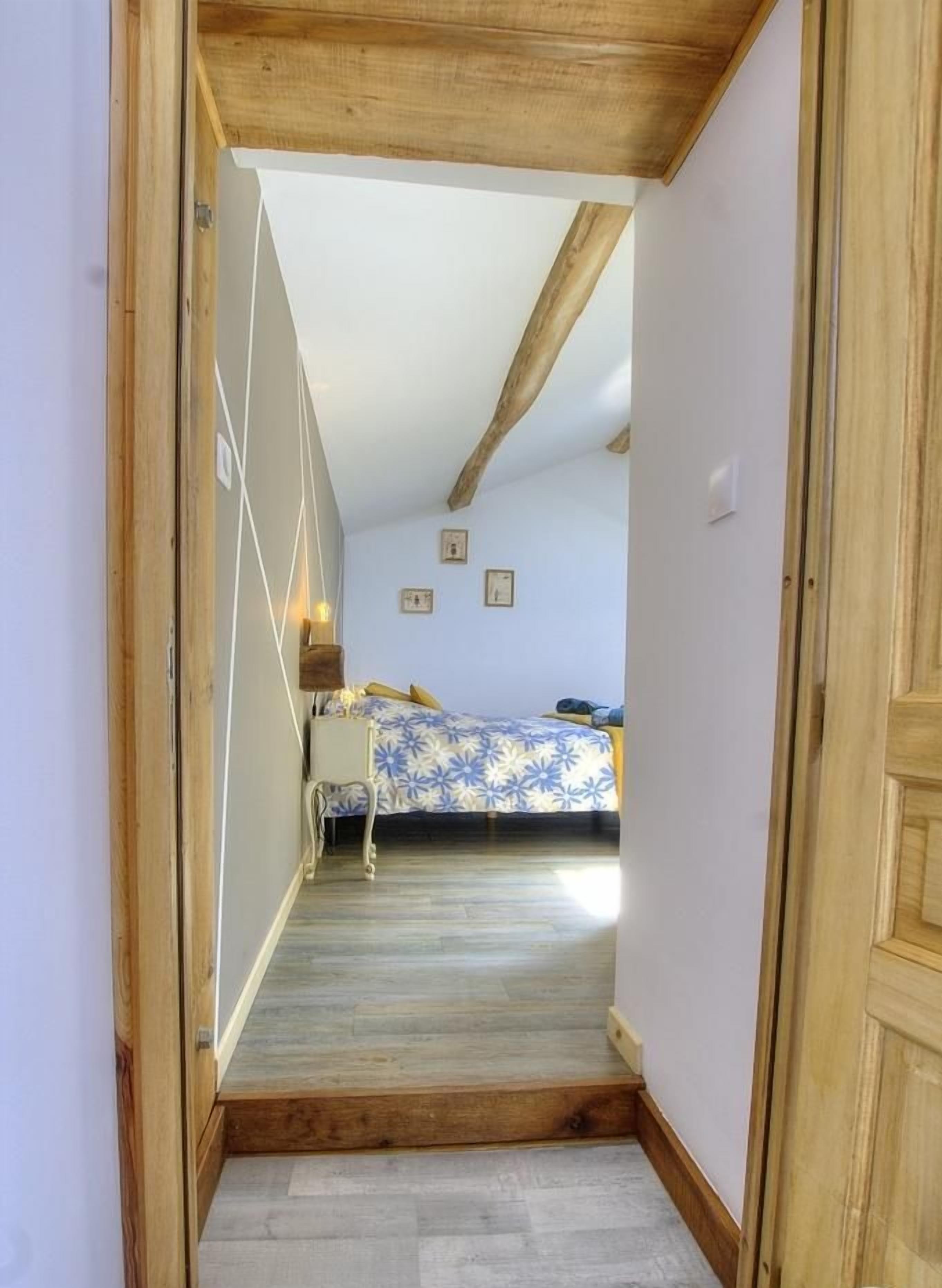 Corredor estreito que dá para um quarto com cama, mesa de cabeceira e luminária na parede.