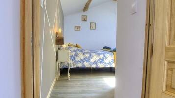 Corredor estreito que dá para um quarto com cama, mesa de cabeceira e luminária na parede.
