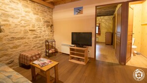 Une pièce chaleureuse avec un mur en pierre, un plafond en bois, un téléviseur posé sur un meuble en bois, une petite table avec un magazine et un banc recouvert d’un plaid.