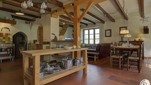 A rustic kitchen with wooden beams, a central island with pots, a dining area with chairs, and a large window providing natural light. - Le Tilleret - Chezery-forens (CHEZERY-FORENS)