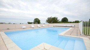 A rectangular swimming pool with a tiled edge, surrounded by a low wall. There are four white lounge chairs aligned along the wall.