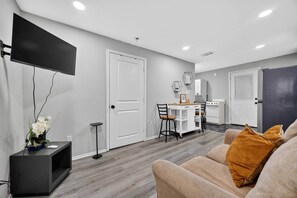 Un salon moderne doté d’un téléviseur à écran plat, d’un canapé agrémenté d’un coussin de couleur moutarde et d’un coin repas avec une cuisine en arrière-plan.