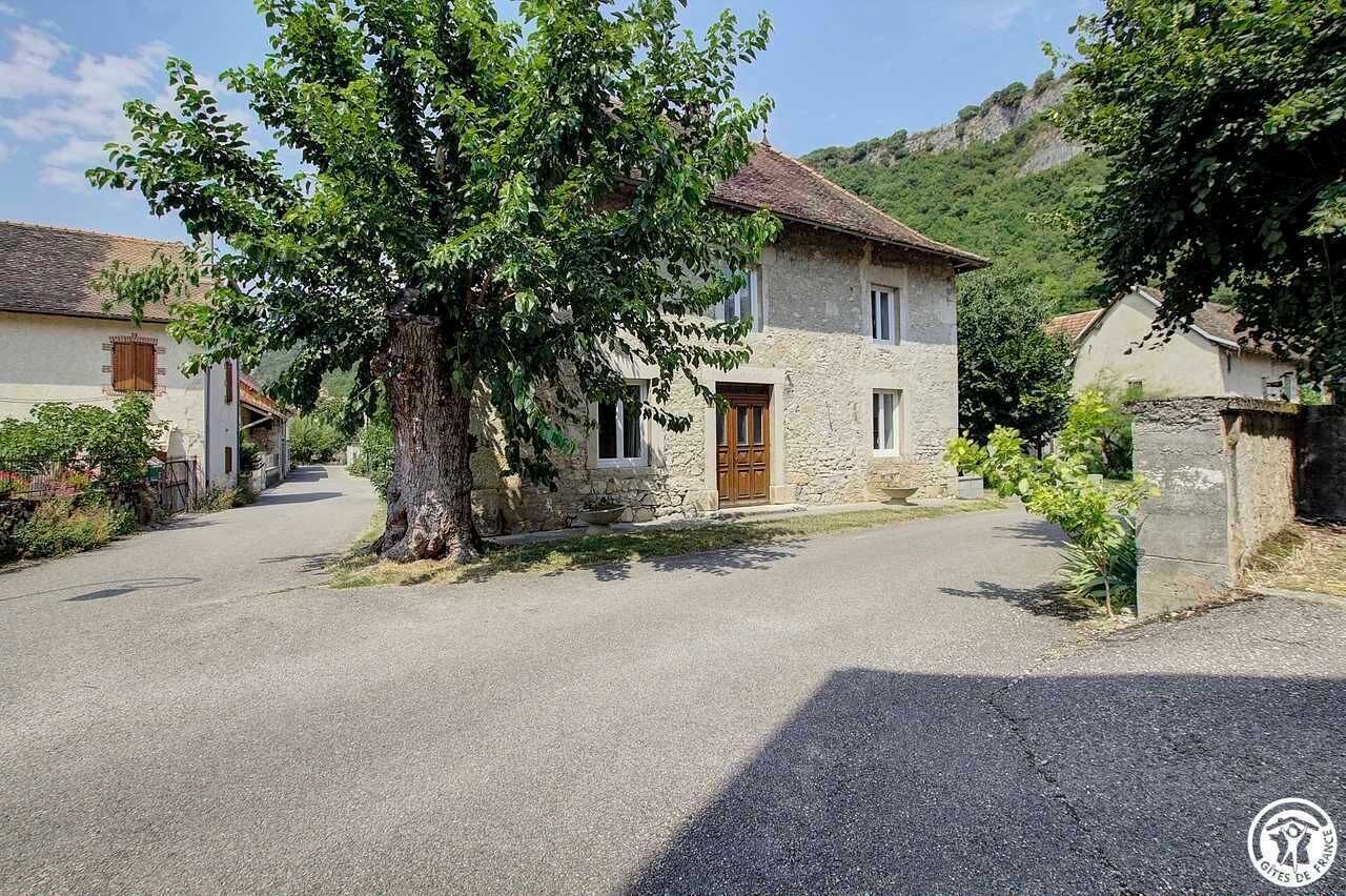 Uma casa de pedra com porta de madeira, um caminho de acesso pavimentado e uma grande árvore em frente.