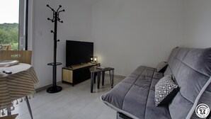 A modern living room with a sofa, a TV, a coat rack, a small table, and a dining area with chairs and a table. - Gîte du Buizin - Vaux-en-bugey (VAUX-EN-BUGEY)