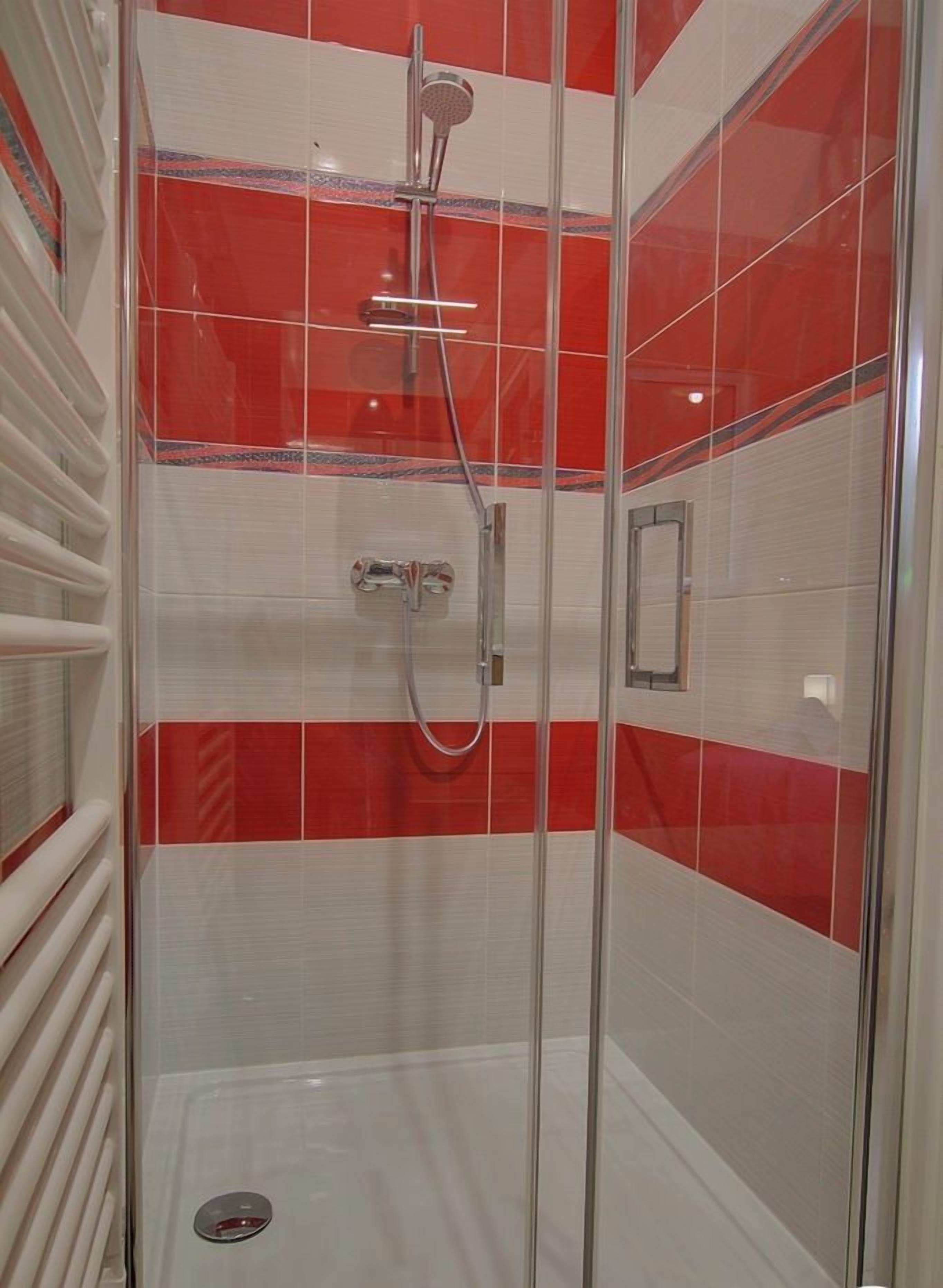 Eine Duschkabine mit Glaswänden, roten und weißen Kacheln, einer Handbrause und einem am Wand montierten Wasserhahn.