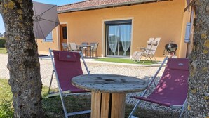Un espace extérieur aménagé avec une table ronde en bois, entourée de deux fauteuils violets, un barbecue et, au fond, un bâtiment avec un toit recouvert de tuiles.