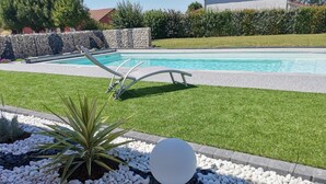 Une piscine bordée d’un mur en pierre et une zone herbeuse agrémentée de plantes et de galets.