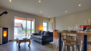 Una sala moderna con chimenea, sofá, mesa de comedor y vista a una casa a través de un ventanal.