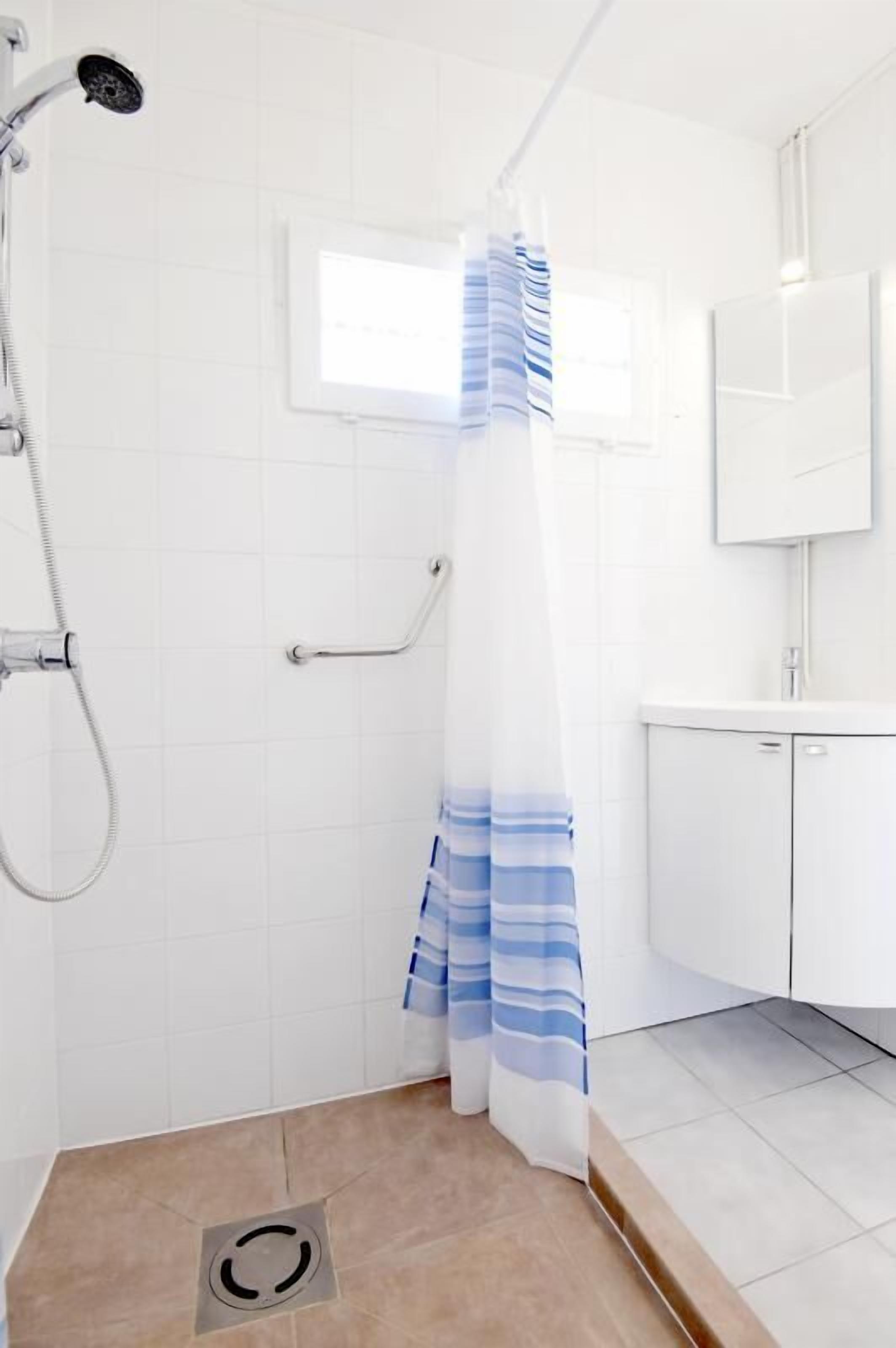 A shower area with a white tiled wall, a window, and a white sink with a cabinet.