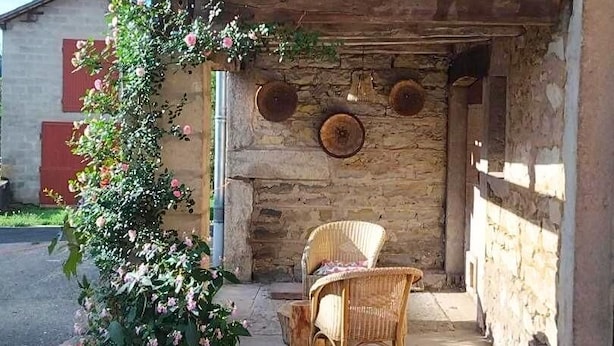 Un edificio de piedra con fachada rústica, una pérgola de madera, sillas de mimbre y una planta en maceta.
