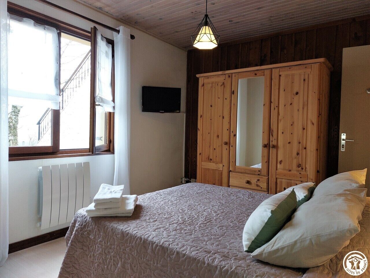 Een slaapkamer met een bed, een houten garderobe, een radiator, een televisie en een raam met gordijnen.