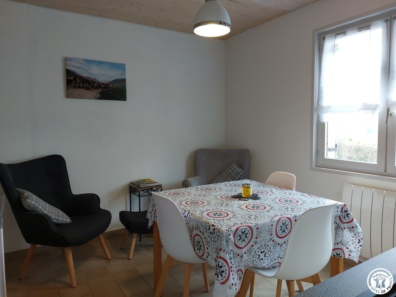 A dining area with a table covered by a patterned tablecloth, two chairs, a black armchair, and a small table with a book and a jar.