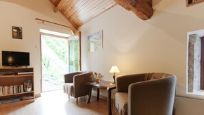 A cozy living room with a wooden ceiling, a television, a bookshelf, two armchairs, a small table with a lamp, and a window with a view of greenery.