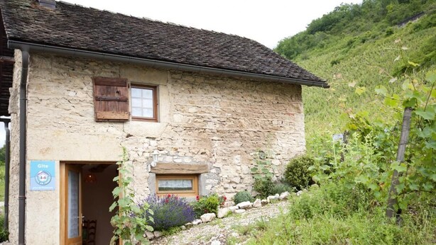 A stone building with a wooden door and window shutters, surrounded by greenery and a hillside.