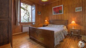 A wooden-paneled bedroom with a bed, two bedside lamps, a window with curtains, and a radiator. - Futin - Outriaz - Gîte (OUTRIAZ)