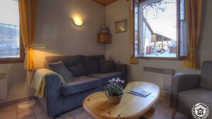 A cozy room with a sofa, a wooden coffee table with a plant, and a window with curtains. - Futin - Outriaz - Gîte (OUTRIAZ)