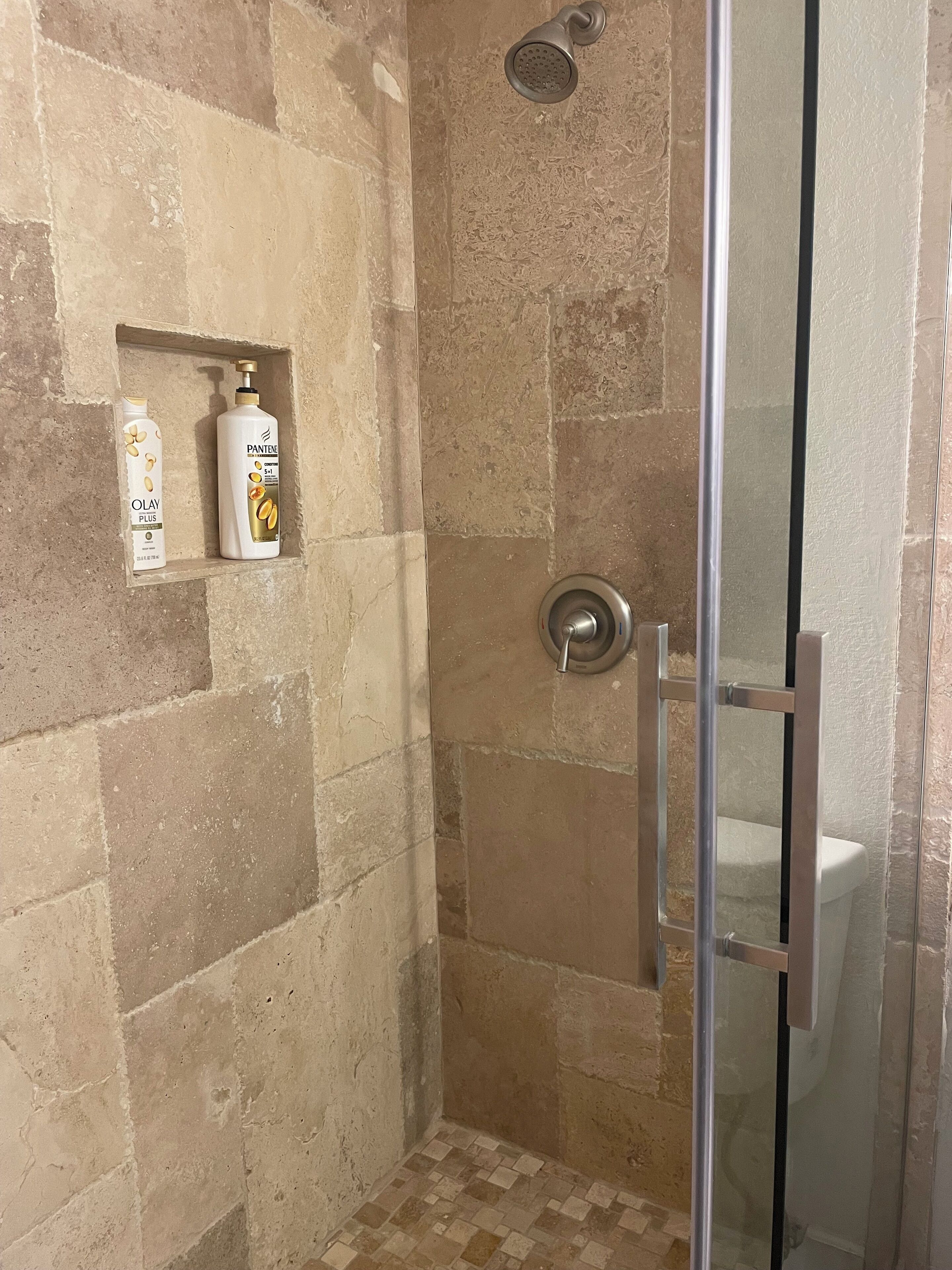 Un área de ducha con puerta de vidrio, azulejos color beige y un estante con botellas de artículos de aseo.