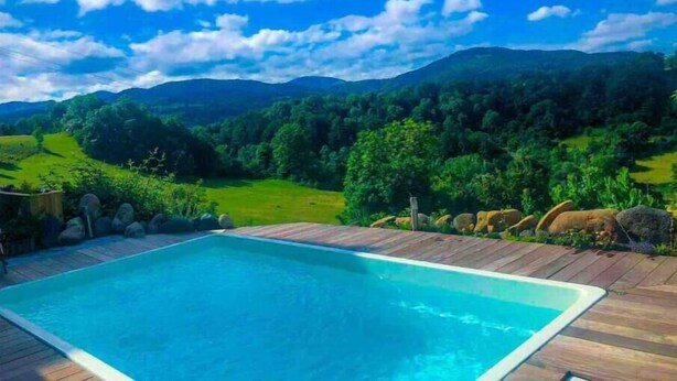 A rectangular swimming pool with a wooden deck, surrounded by a lush green landscape and mountains in the background. - Beaume d’ataraxie - Beautiful View Room (Apt. 4) (Solignac-sur-Loire)