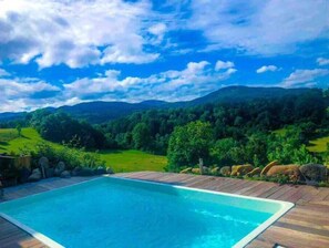 A rectangular swimming pool with a wooden deck, surrounded by a lush green landscape and mountains in the background. - Beaume d’ataraxie - Beautiful View Room (Apt. 4) (Solignac-sur-Loire)