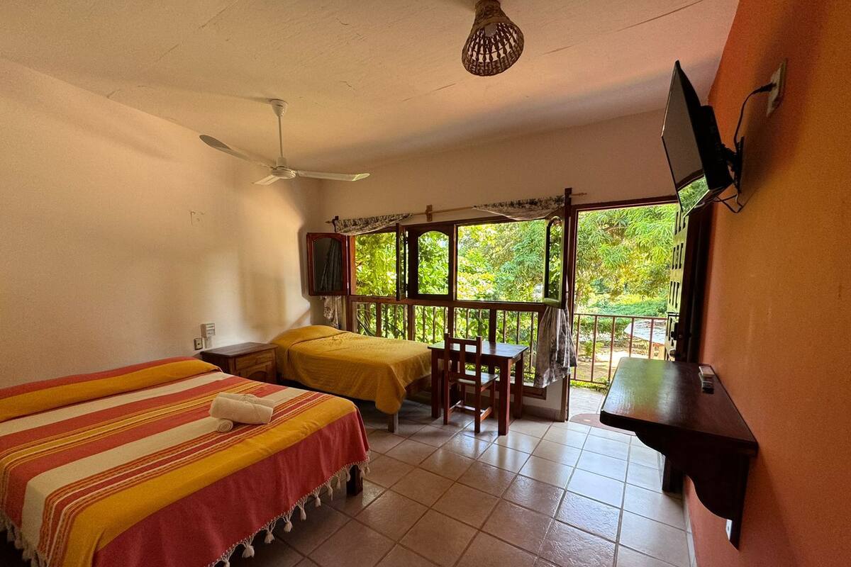 A room with two beds, a table, chairs, a TV, and a balcony with a view of greenery.