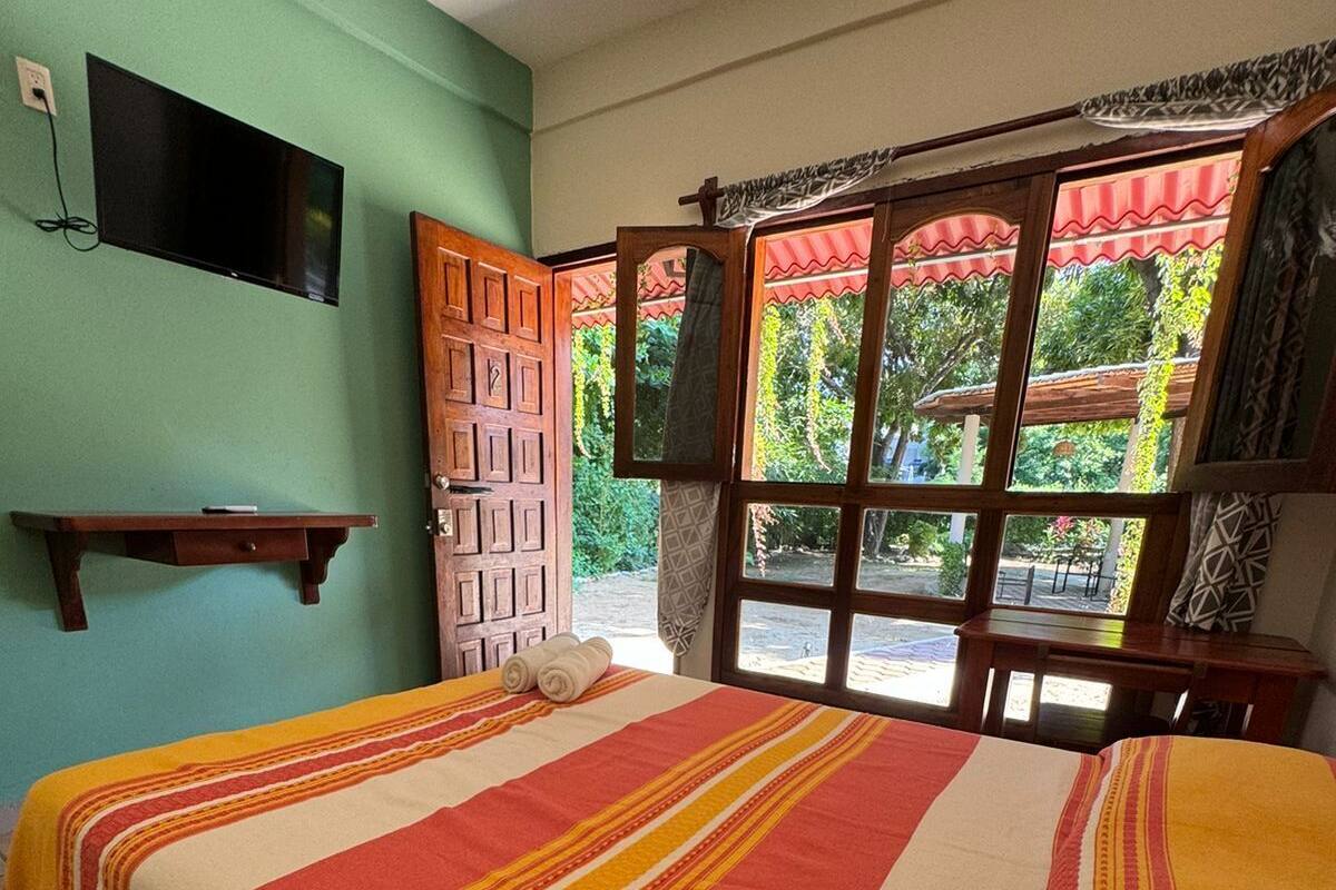 A room with a bed, a TV mounted on the wall, a window with a view of greenery, and a wooden door.