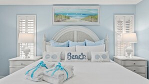 A bedroom with a large bed, two bedside tables, a headboard with a painting, and a beach-themed pillow. - Sunshine House! 3BR 2BA Sleeps 9! Your Perfect Retreat for Comfort and Style (Panama City Beach)