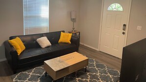 A living room with a black leather sofa, two yellow cushions, a wooden coffee table, a patterned rug, a lamp, and a white door with a window. - Cozy two bedroom home minutes from downtown (Houston)